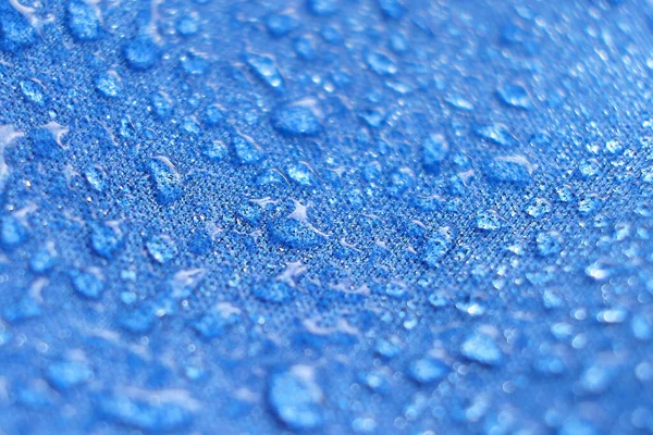 Wasser tropft auf den Stoff. Regenwassertropfen auf blauem, faserdichtem Gewebe. Wassertropfen ziehen sich über ein wasserdichtes Tuch. Blauer Hintergrund. — Stockfoto