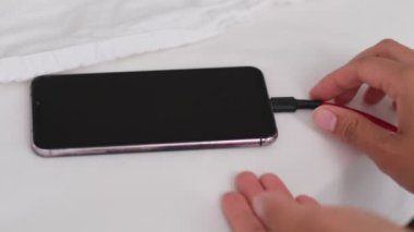 Akıllı telefon kırışık beyaz çarşaflar içinde yatakta uzanıyor, sabahları şarj oluyor, kapanıyor. Yatak odası arka planı. Bir telefon cihazını başıboş bırakmak, soyut kavramları şarj etmek.