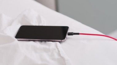 Akıllı telefon kırışık beyaz çarşaflar içinde yatakta uzanıyor, sabahları şarj oluyor, kapanıyor. Yatak odası arka planı. Bir telefon cihazını başıboş bırakmak, soyut kavramları şarj etmek.