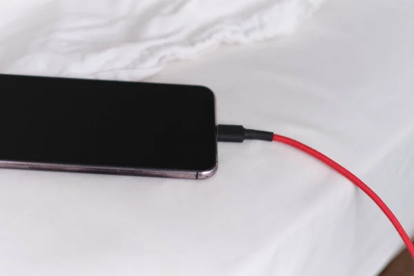 Teléfono inteligente se está cargando en la cama y acostado en tela blanca. Adicto al teléfono inteligente y concepto de estilo de vida saludable. Cable USB conectado con un teléfono móvil blanco — Foto de Stock