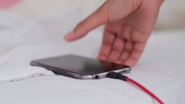 Akıllı telefon yatakta şarj oluyor ve beyaz kumaşın üzerinde yatıyor. Akıllı telefon bağımlısı ve sağlıklı yaşam tarzı konsepti. USB kablosu beyaz bir cep telefonuna bağlandı