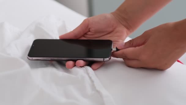スマートフォンはベッドに充電され、白い布に横たわっています。スマートフォン中毒者と健康的なライフスタイルの概念。白い携帯電話に接続されたUSBケーブル — ストック動画