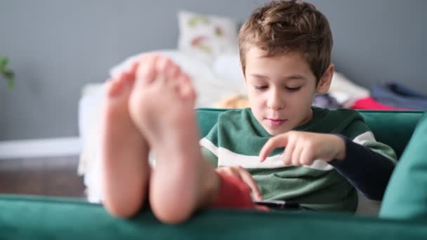 Niño alegre sonriendo mientras está sentado en el sofá y usando la tableta en casa. Tecnología moderna para niños y educación. pequeño niño mirando almohadilla táctil en casa. — Vídeos de Stock