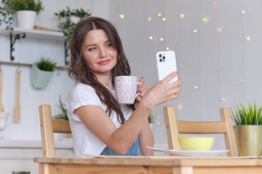 Mutfakta elinde kahve tutarak gülümseyen bir kadın. Online mağazalardan yiyecek sipariş etmek. çevrimiçi bir uygulama aracılığıyla yiyecek satın almak