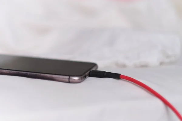 Teléfono inteligente se está cargando en la cama y acostado en tela blanca. Adicto al teléfono inteligente y concepto de estilo de vida saludable. Cable USB conectado con un teléfono móvil blanco — Foto de Stock