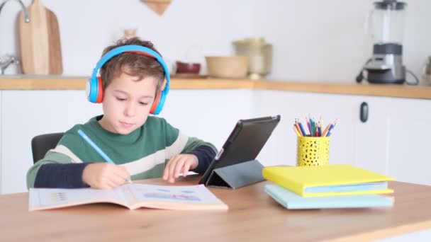 Kleine jongen gebruikt een laptop om een videogesprek te voeren met zijn leraar. jongen die op afstand online studeert schrijft de lessen die de leraar vertelde in zijn notitieboekje. Concept afstandsonderwijs. — Stockvideo