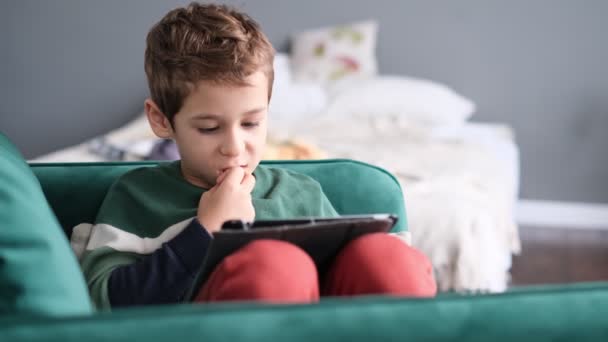 Niño alegre sonriendo mientras está sentado en el sofá y usando la tableta en casa. Tecnología moderna para niños y educación. pequeño niño mirando almohadilla táctil en casa. — Vídeos de Stock