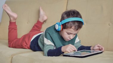 Kanepede yatıp tablet kullanırken gülümseyen neşeli küçük çocuk. Modern çocuk ve eğitim teknolojisi. Küçük çocuk evdeki dokunmatik yere bakıyor..