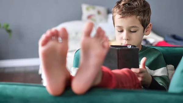 Χαρούμενο αγοράκι που χαμογελάει ενώ κάθεται στον καναπέ και χρησιμοποιεί tablet στο σπίτι. Σύγχρονη παιδική και εκπαιδευτική τεχνολογία. μικρό αγόρι που κοιτάζει το σημειωματάριο στο σπίτι. — Φωτογραφία Αρχείου