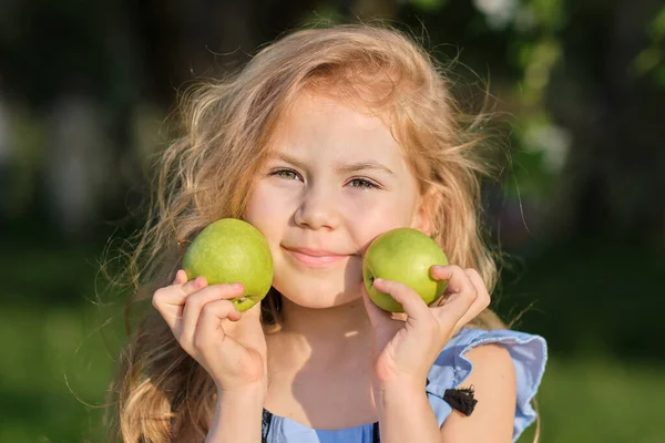 La niñita sonríe a la cámara. chica bonita posando para una foto y sosteniendo dos manzanas verdes frescas en sus manos — Foto de Stock