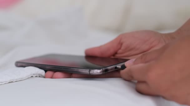 Смартфон заряжается на кровати и лежит на белой ткани. Зависимость от смартфонов и здоровый образ жизни. Кабель USB, соединенный с белым мобильным телефоном — стоковое видео