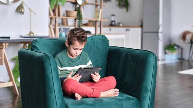 Kanepede oturup tablet kullanırken gülümseyen neşeli küçük çocuk. Modern çocuk ve eğitim teknolojisi. Küçük çocuk evdeki dokunmatik yere bakıyor..
