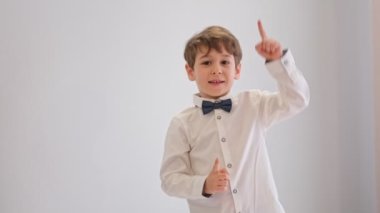 Beyaz arka planda, parmakları yukarı doğru gösteren sevimli çocuk şaşırdı. Papyonlu beyaz gömlekli çocuk, genç beyefendi.