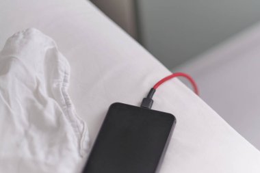 Akıllı telefon yatakta şarj oluyor ve beyaz kumaşın üzerinde yatıyor. Akıllı telefon bağımlısı ve sağlıklı yaşam tarzı konsepti. USB kablosu beyaz bir cep telefonuna bağlandı