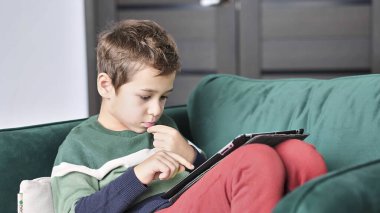 Kanepede oturup tablet kullanırken gülümseyen neşeli küçük çocuk. Modern çocuk ve eğitim teknolojisi. Küçük çocuk evdeki dokunmatik yere bakıyor..