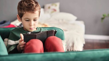 Kanepede oturup tablet kullanırken gülümseyen neşeli küçük çocuk. Modern çocuk ve eğitim teknolojisi. Küçük çocuk evdeki dokunmatik yere bakıyor..