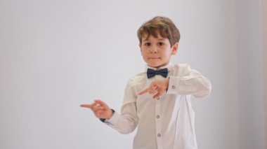 Genç ve tapılası çocuk beyaz arka planda önce solu, sonra sağı işaret ediyor. Papyonlu beyaz gömlekli çocuk, genç beyefendi. Çocuk dans ediyor ve sola bakıyor.