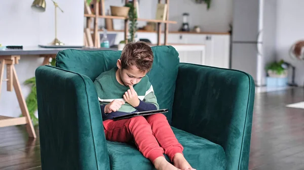 Kanepede oturup tablet kullanırken gülümseyen neşeli küçük çocuk. Modern çocuk ve eğitim teknolojisi. Küçük çocuk evdeki dokunmatik yere bakıyor..