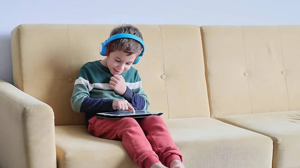 Fröhlicher kleiner Junge lächelt, während er auf dem Sofa sitzt und zu Hause Tabletten benutzt. Moderne Kinder- und Bildungstechnologie. Kleiner Junge schaut zu Hause auf Touchpad. — Stockfoto