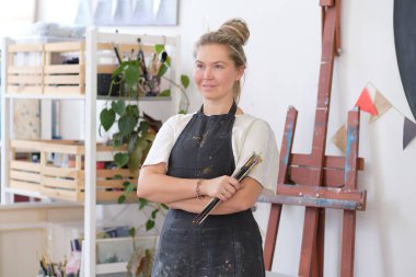 Kapalı alanda fırçayla poz veren yetenekli bir kadın sanatçı. Fırça tutarken kollarını kavuşturur, gülümseyerek kameraya bakar. Geniş tuvalli otantik yaratıcı stüdyo.