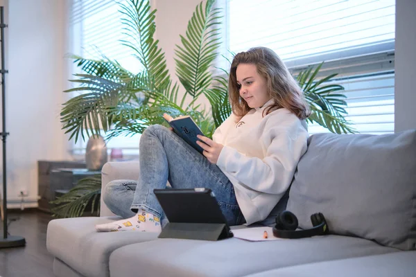 A lány leül a kanapéra, és könyvet olvas, egy tablettát és fülhallgatót hever mellette. modern tinédzser otthon. tini irodalom koncepció. — Stock Fotó