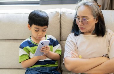 Asyalı bir çocuk akıllı telefon kullanırken, hayal kırıklığına uğramış bir anne onu izliyor, evdeki kanepede oturuyor. Telefon bağımlılığı kavramı.
