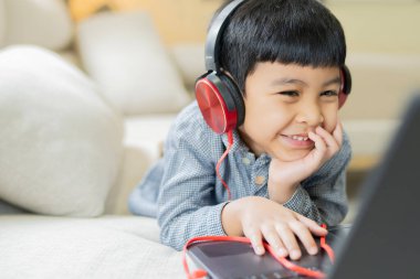 Evdeki online derste öğretmeniyle iletişim kurarken bilgisayar ekranına bakan kulaklık takan neşeli Asyalı küçük çocuk.