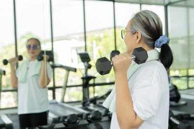 Ağırlık çalışan, ağırlık kaldıran spor salonundaki aynada kendine bakan neşeli, olgun Asyalı kadın. Seçici odak.