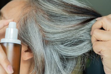 Orta yaşlı, gri saçlı, elinde kahverengi bir şişeyle saçlarını uzatan bir kadın. Saç spreyi, kozmetik ürünler konsepti.