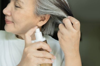 Olgun, gümüş saçlı Asyalı bir kadın elinde saç serumu tutarken kendine güvenerek gülümsüyor. Seçici odaklanma. Saç tedavisi kavramı.