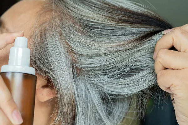Orta yaşlı, gri saçlı, elinde kahverengi bir şişeyle saçlarını uzatan bir kadın. Saç spreyi, kozmetik ürünler konsepti.