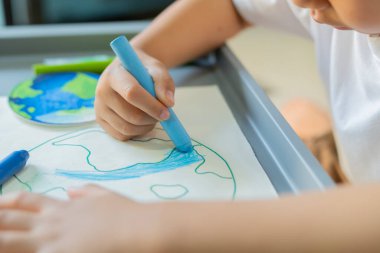 Evdeki masada oturan bir çocuk, beyaz kağıda çok renkli kalemler çizen bir dünya haritası çiziyor. Barış ve Toprak Günü kavramı.