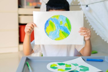 Küçük Asyalı çocuk oyun odasında eko-temalı çizimler tutuyor, çevresel farkındalığı ve yaratıcılığı teşvik ediyor. Dünya Günü konsepti