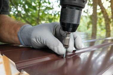 Metal bir çatıya vida sıkıştırmak için elektrikli tornavida kullanan bir adam. Gri iş eldivenli bir elin, tornavidayla kendini beceren bir tornavidayı metal çatı profiline sokması.