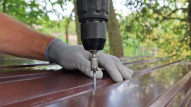 Kendini beceren bir vidayı metal bir levhaya vidalamak. Gri eldivenli bir zanaatkarın elinde bir tornavida var. Kahverengi metal profil profiline elektrikli vidayla vida takıyor.