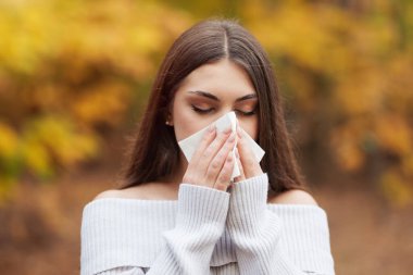 Kağıt mendil kullanan, burnu akan genç bir kadın. Bir kadın burnunu siliyor, yüzünü bir peçeteyle örtüyor, sonbahar ormanının arka planına karşı, mevsimsel bir sonbahar soğuğunu resmediyor.