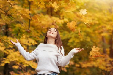 Sarı akçaağaç yapraklarını sonbahar parkında havaya atan mutlu kız. Elleri havada, gülümseyen, düşen sarı yaprakların hareketlerini izleyen genç bir kadın, sonbahar mevsiminin güzelliği.