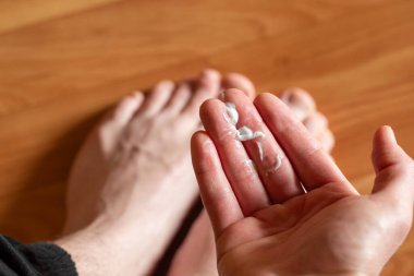Nemlendirici krem sürüyorum. Bir adamın eline az miktarda beyaz kremle yakın çekim, tahta zeminde ayaklara ve bacaklara uygulanmaya hazırlanmak, bakım, nemlendirici, ayak bakımı