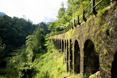 Parlak, doğal güneş ışığıyla aydınlatılan yeşil ormanlı bir dağ bölgesinde uzanan kemerli antik bir su kemerinin manzarası, seyahat ve doğa temalı kullanım için ideal barışçıl ve pitoresk bir atmosfer yaratıyor..