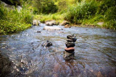 Berrak akan su ve yemyeşil bitki örtüsüyle çevrili dengeli taşlarla dolu huzurlu bir nehir açık hava, doğa ve rahatlama temaları için uygun sakin bir doğal manzara yaratıyor..