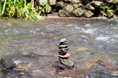 Akan bir derede dengelenmiş küçük taşlar yığını ve arka planda yeşil bitkiler ve kayalar açık hava ya da doğa temalı projeler için sükunet ve doğal güzelliği çağrıştırıyor..