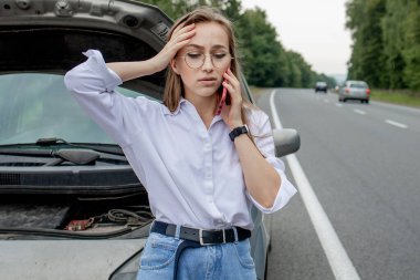 Young woman standing near broken down car with popped up hood having trouble with her vehicle. Waiting for help tow truck or technical support. A woman calls the service center. clipart