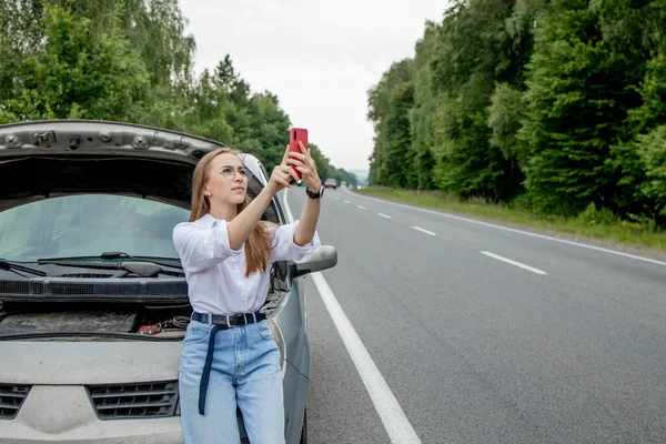 壊れた車の近くに立っている若い女性は 彼女の車に問題があるフードをポップアップしました トラックや技術サポートを牽引するのを待っています 女性がサービスセンターに電話して — ストック写真