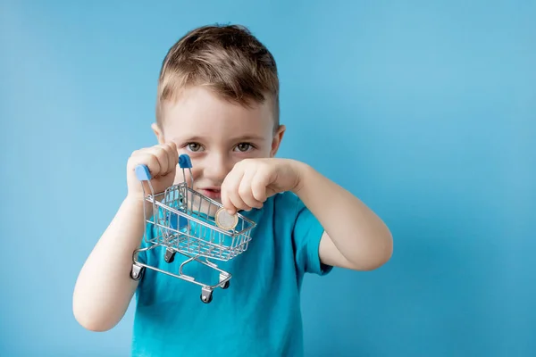 身穿蓝色T恤的男孩拿着蓝色背景的购物车和硬币 购物和销售概念 — 图库照片