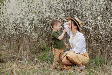 Mutlu anne ve oğul birlikte eğleniyor. Anne oğluna nazikçe sarılıyor. Arka planda beyaz çiçekler açar. Anneler Günü..