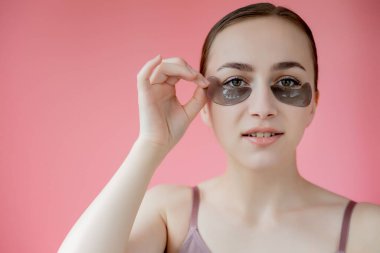 Baş portresi. Göz altı nemlendirici maskeli gülümseyen genç bir kadın. Kameraya bakıp cilt bakımının keyfini çıkarıyor.