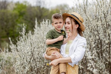 Mutlu anne ve oğul birlikte eğleniyor. Anne oğluna nazikçe sarılıyor. Arka planda beyaz çiçekler açar. Anneler Günü..