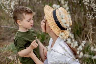 Mutlu anne ve oğul birlikte eğleniyor. Anne oğluna nazikçe sarılıyor. Arka planda beyaz çiçekler açar. Anneler Günü..