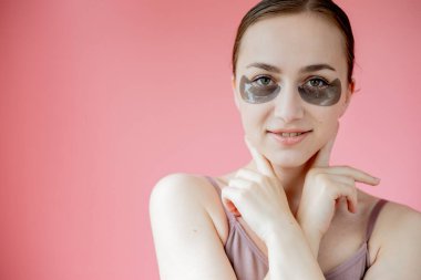 Baş portresi. Göz altı nemlendirici maskeli gülümseyen genç bir kadın. Kameraya bakıp cilt bakımının keyfini çıkarıyor..