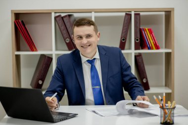 Tipik çalışma günü. Ofisinde oturan ve dizüstü bilgisayarına bakan kendinden emin genç bir iş adamının fotoğrafını çek..